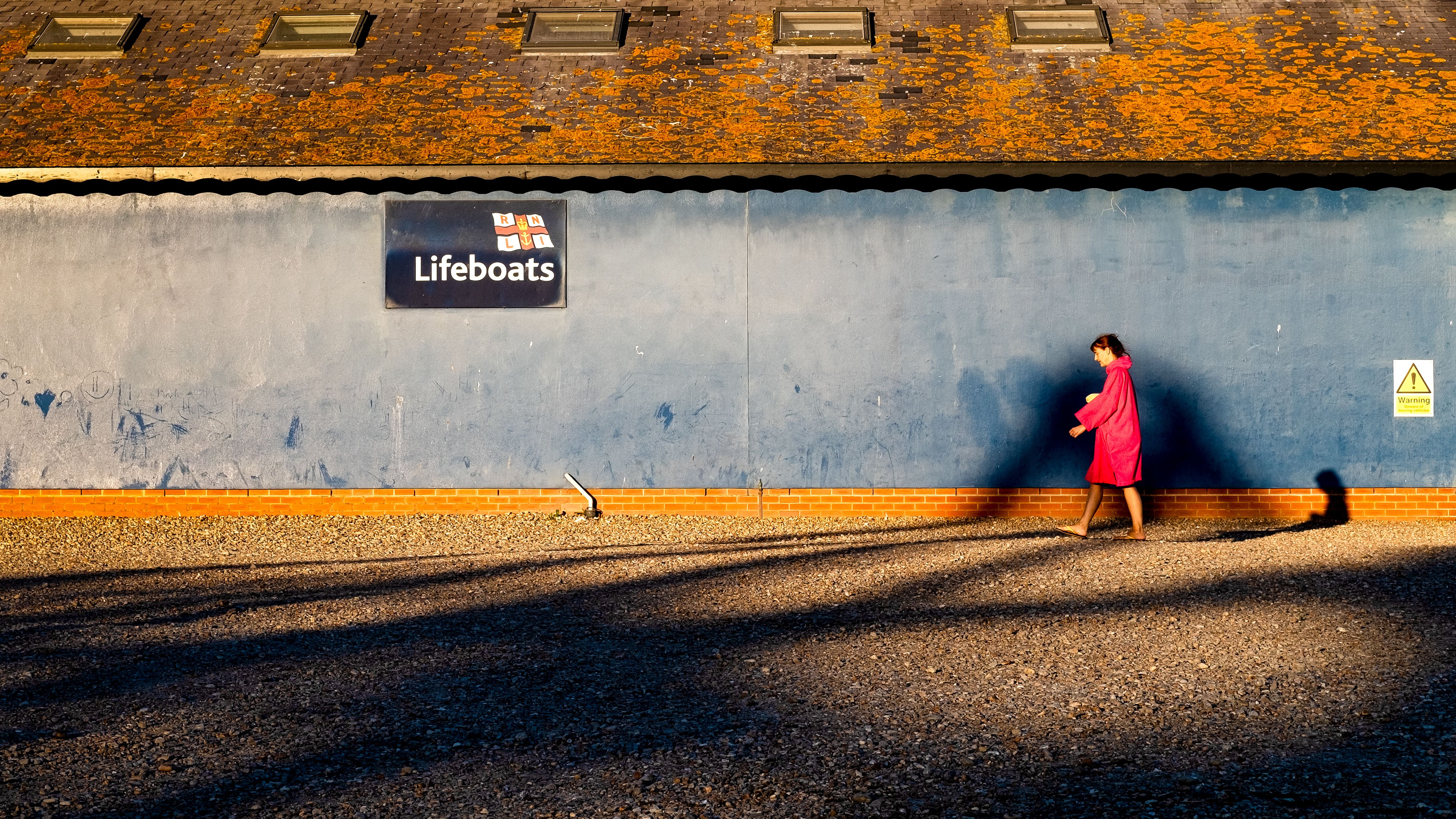 Vrouw in rode jas loopt langs een oranje verlichte muur met een Lifeboats-bord, lange schaduwen