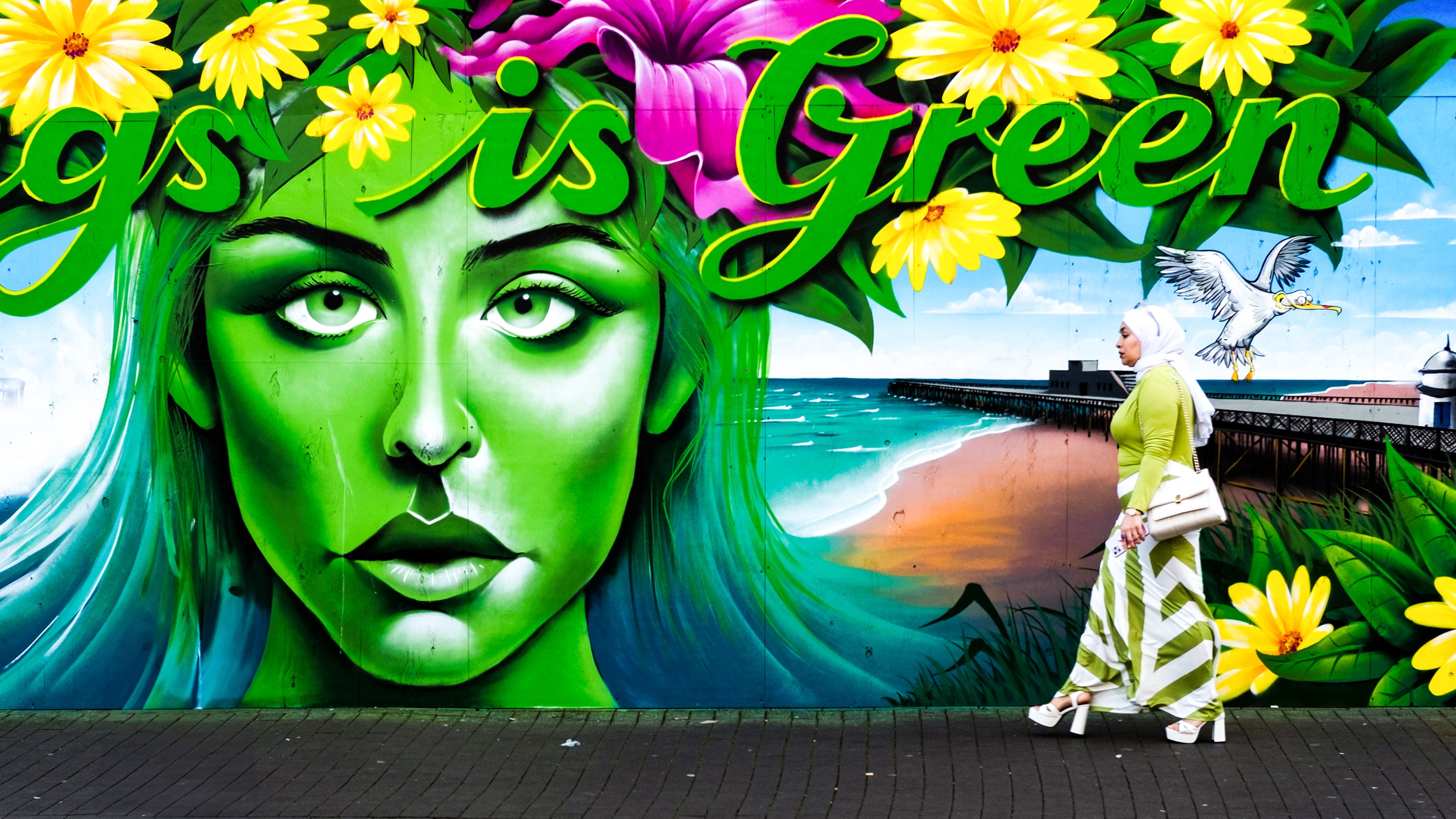 Muurschildering van een groen vrouwengezicht met bloemen, een vrouw loopt voorbij
