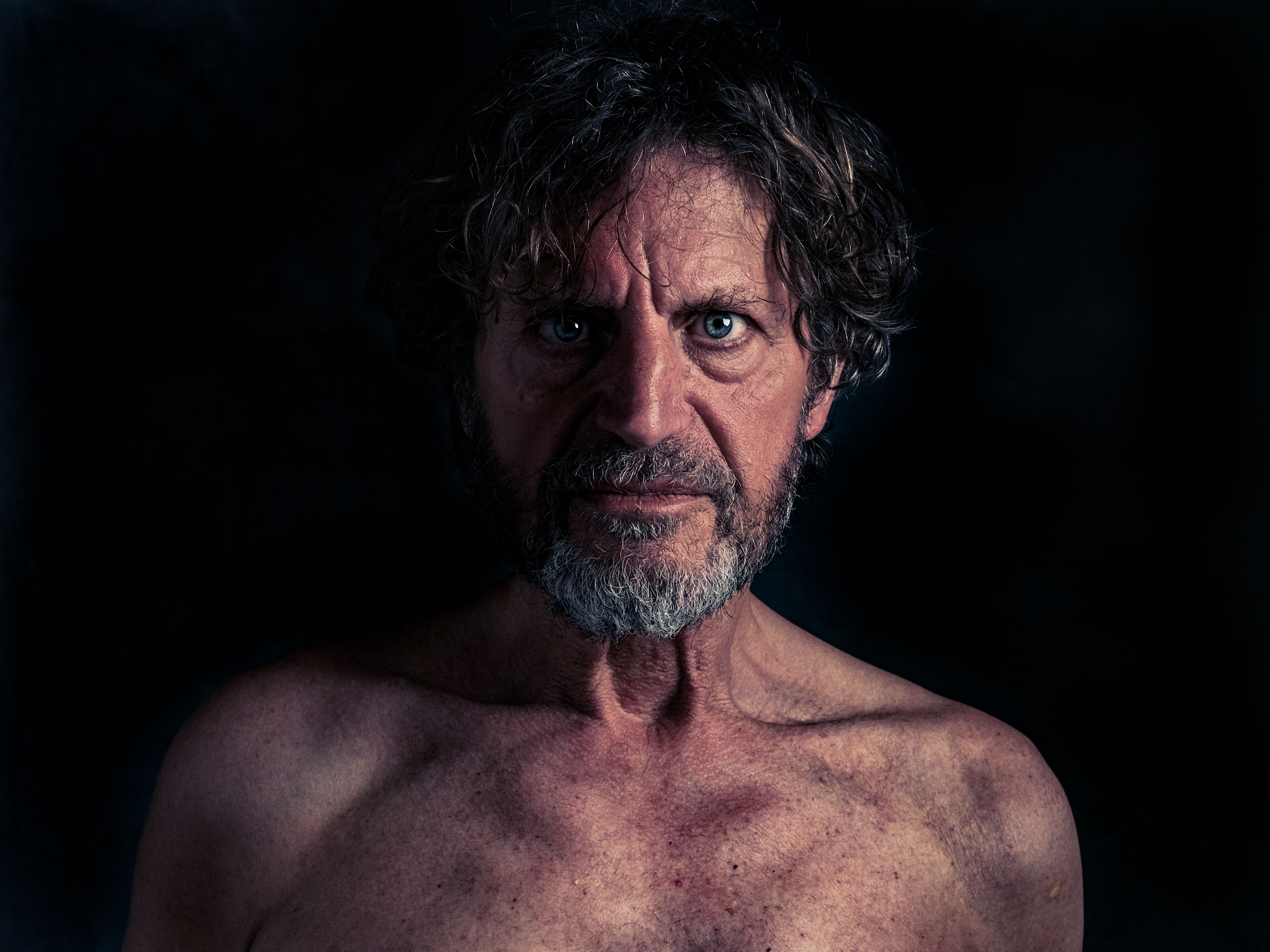 Portret van een oudere man met ontbloot bovenlijf en indringende blik, tegen een donkere achtergrond