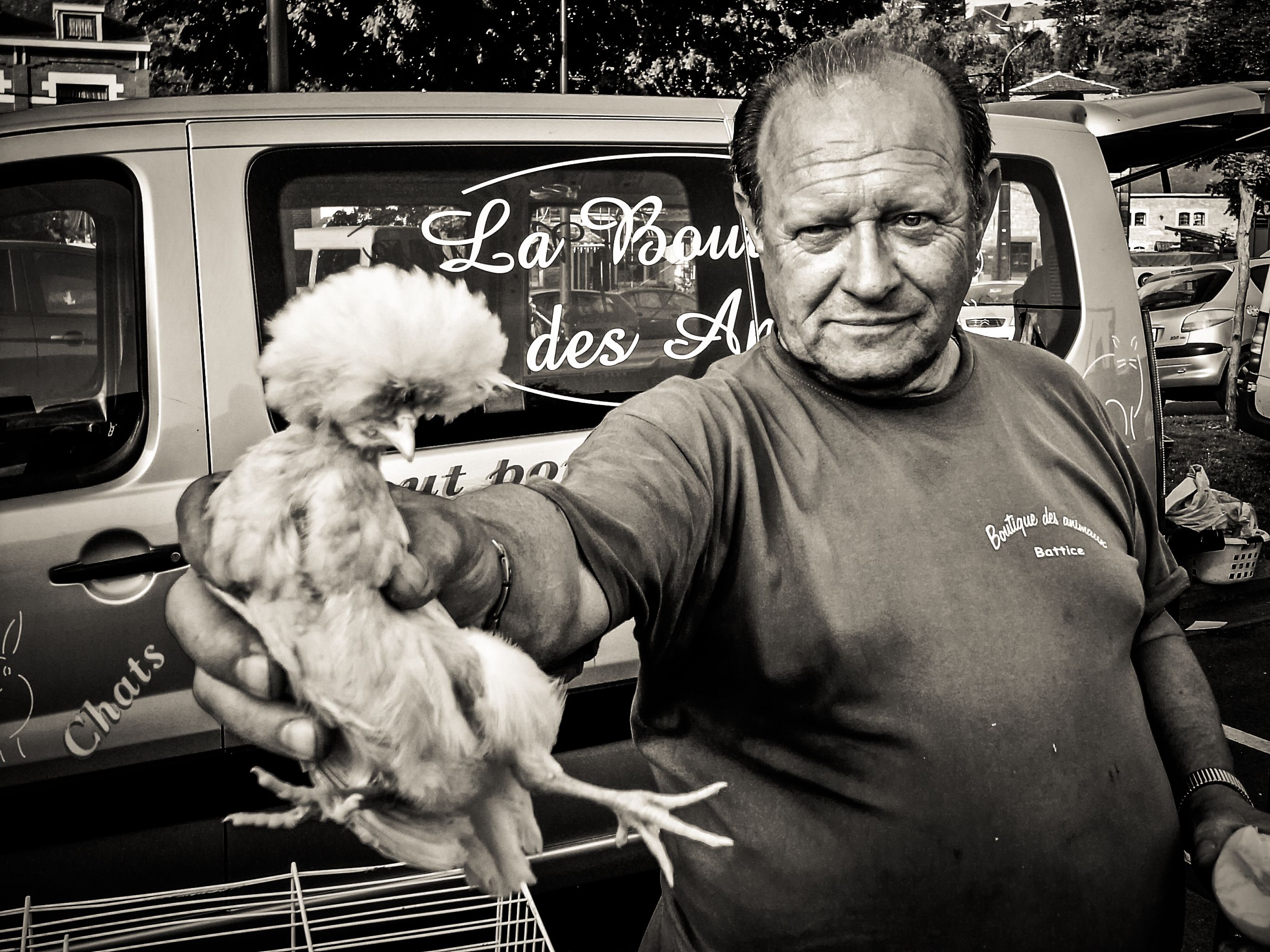 Man op een markt in Esneux houdt lachend een witte kip omhoog