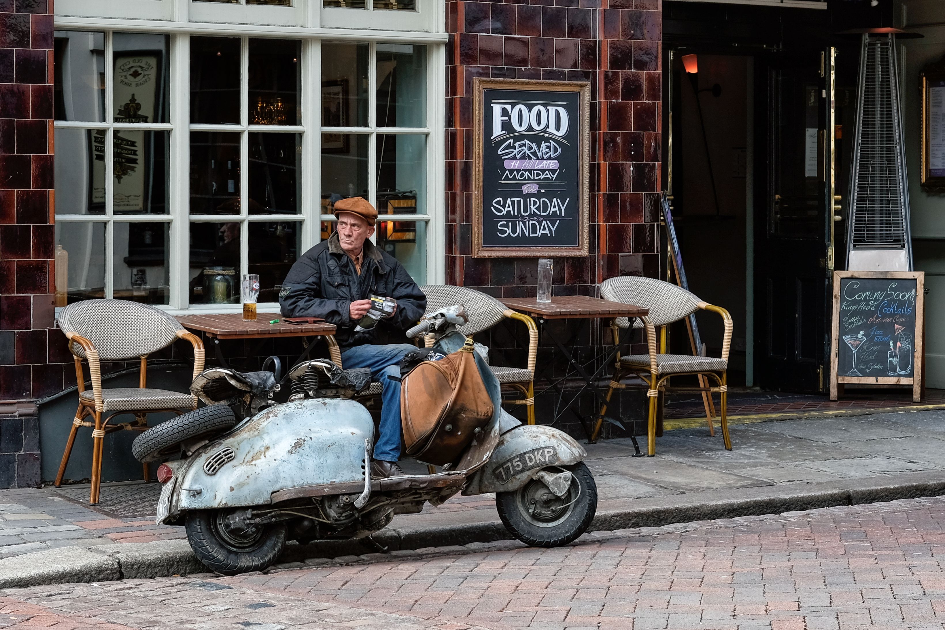 Man op een terras naast een oude scooter voor een tegelgevel